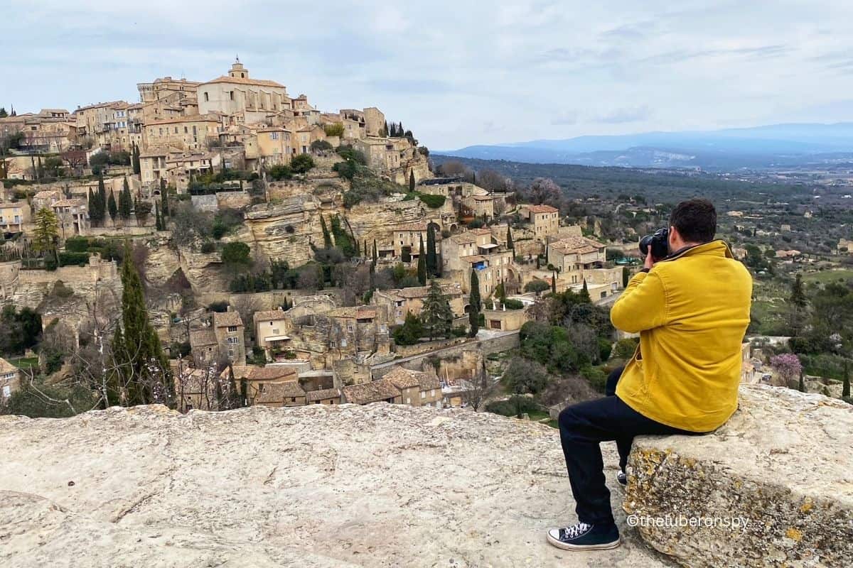 Man in a yellow jacket taking a photo with his camera of the perched village of Gordes