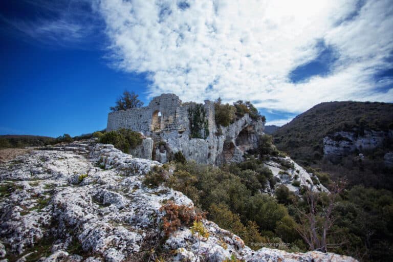 Ruins of the Fort de Buoux perched atop a rocky cliff under a vivid blue sky with wispy clouds.