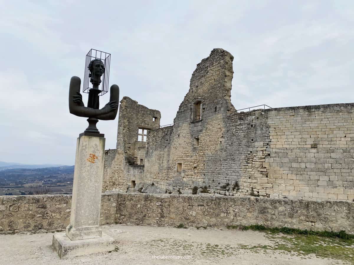 Modern black metal sculpture featuring a caged head atop crossed arms stands before the ruined walls of Chateau de Sade in Lacoste.