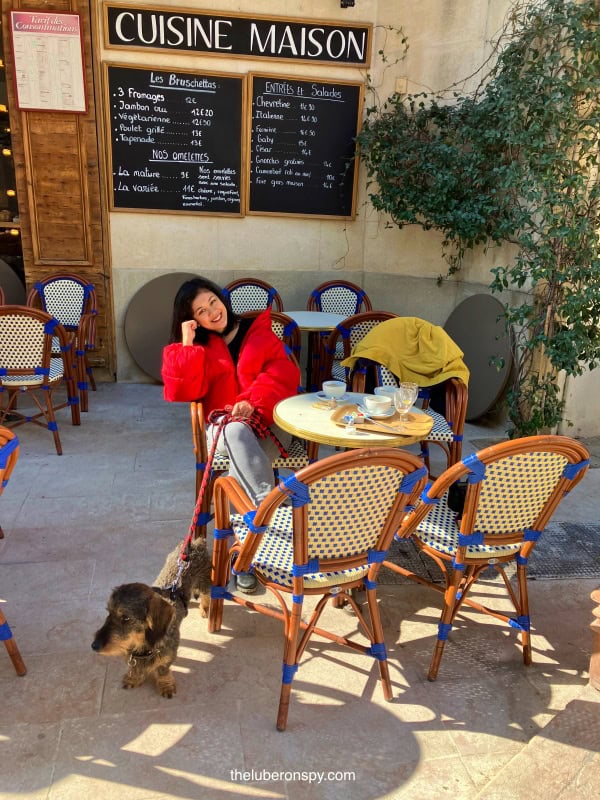 Smiling woman (this writer) in a red jacket sits at a café terrace in Luberon, with her dog by her side, under a 'Cuisine Maison' sign.