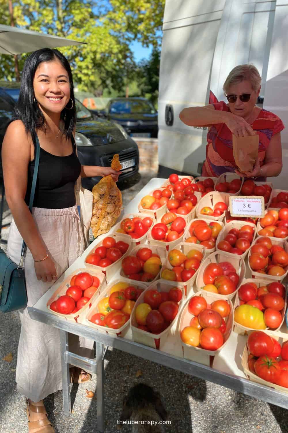 Smiling Jenny (the writer) at a market stall holding tomatoes in a Luberon market, with baskets of fresh tomatoes on display.