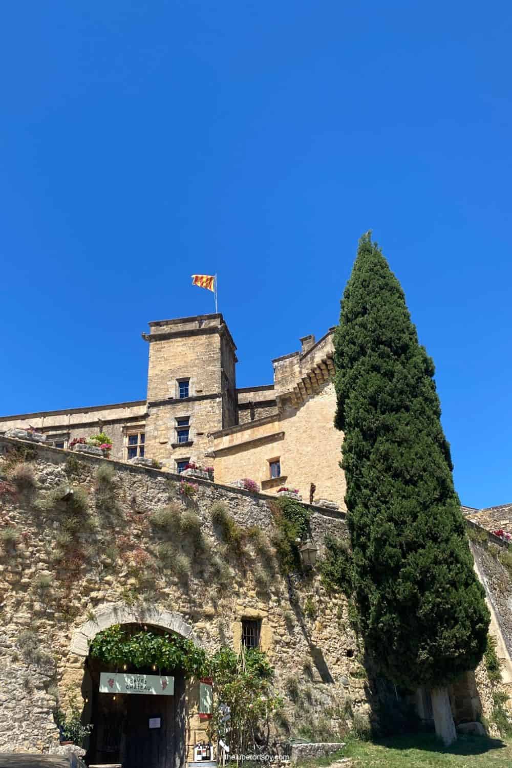 Historic Lourmarin Castle in France with a colorful flag atop its tower and a quaint wine shop nestled below, under a clear blue sky.