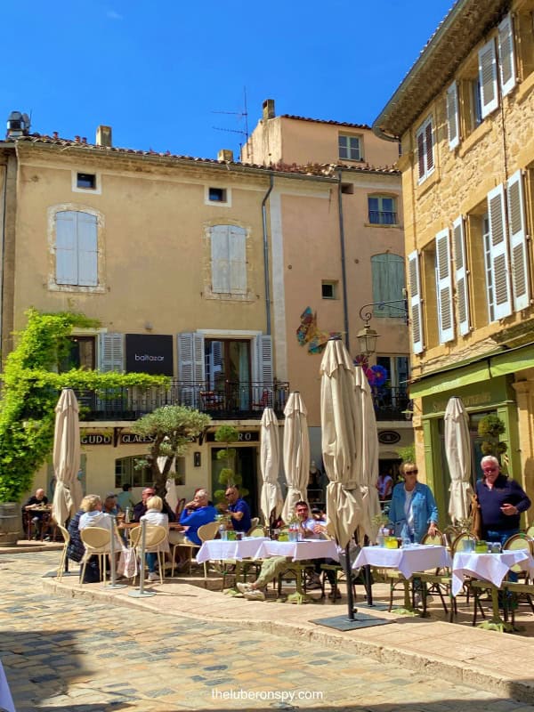 Diners enjoy lunch at a charming outdoor café in a sunlit cobblestone square in Luberon, surrounded by picturesque buildings.