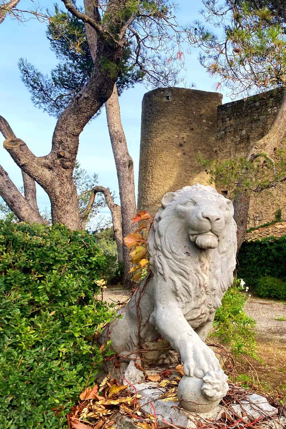 Stone lion statue in Menerbes, France, with autumn leaves, standing proudly near ancient stone walls and trees.