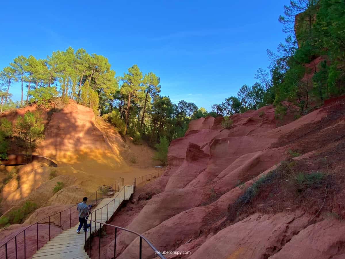 A man walks along a wooden path winding through the stunning red and yellow ocher cliffs of the Ocher Trail in Roussillon, France.