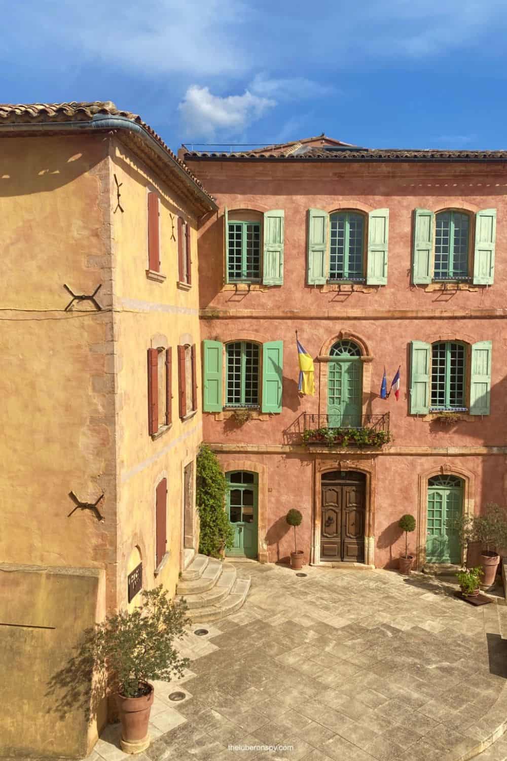 Charming pastel buildings with colorful shutters in Roussillon, bask in the sunlight under a bright blue sky.