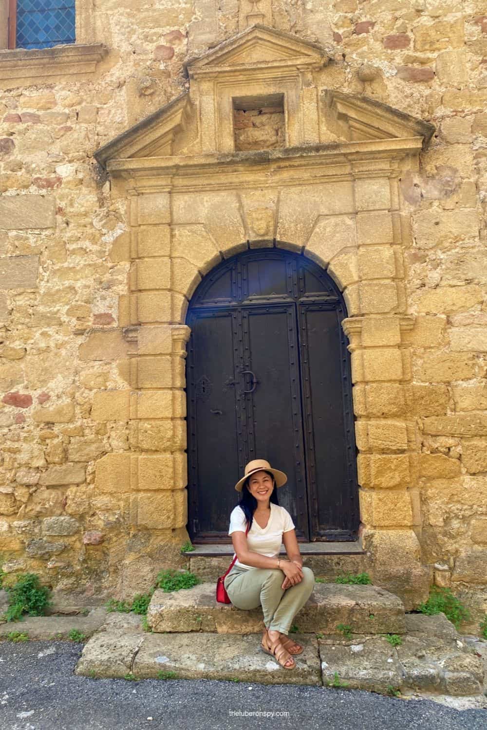 Smiling woman (the Luberon Spy writer) in a hat sits on stone steps in front of a historic, arched doorway in Ansouis, France.