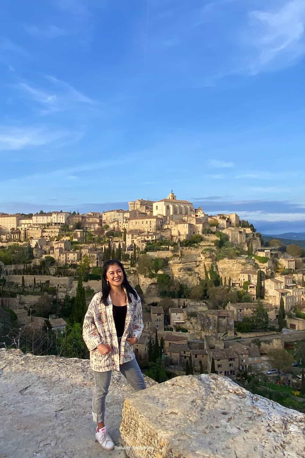 Smiling woman (the Luberon Spy writer) stands on a cliff with the picturesque village of Gordes, France, in the background under a clear blue sky.
