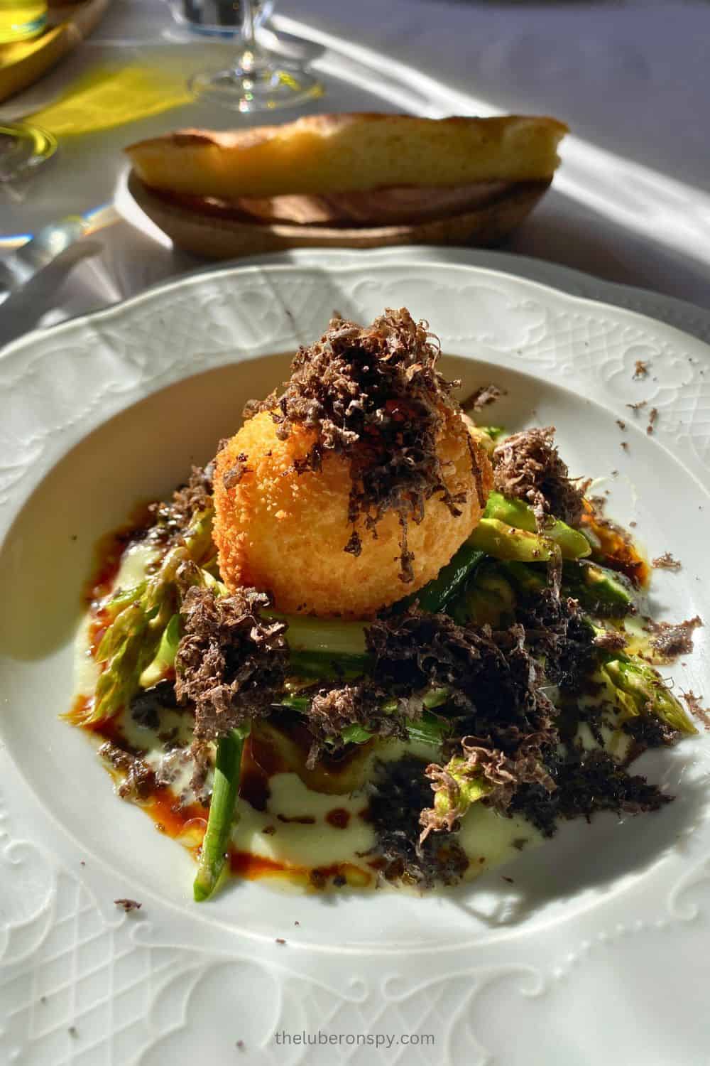 A gourmet dish featuring a crispy fried ball garnished with generous truffle shavings, served on a bed of asparagus with a rich, creamy sauce. A slice of bread and a glass of wine accompany the meal, highlighting the culinary elegance.
