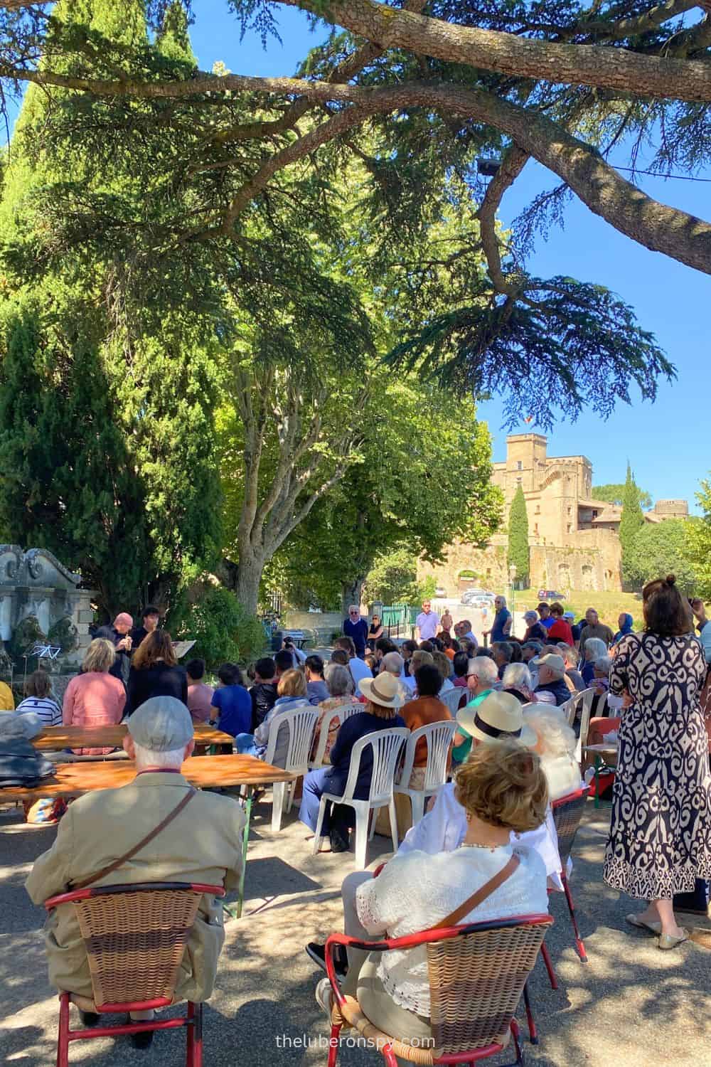 People enjoying a free outdoor music concert in Lourmarin, with a backdrop of lush greenery and the castle.