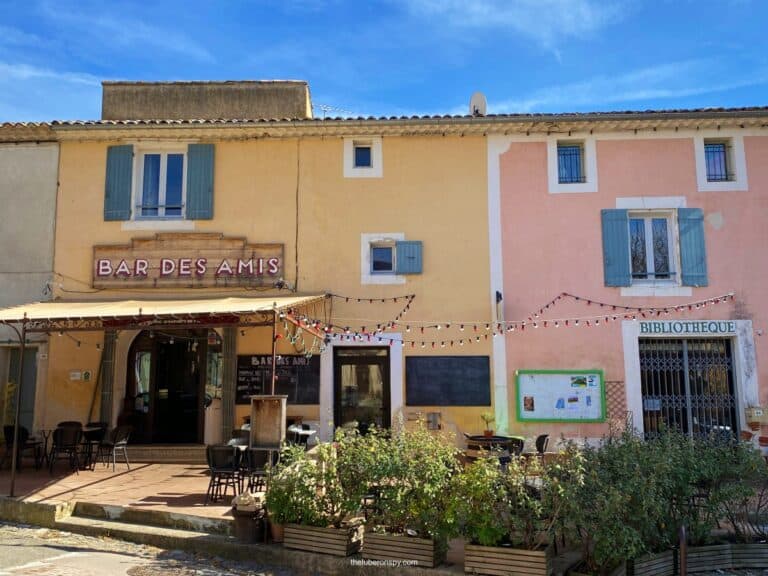 Bar des Amis in Villars, featuring a pastel-colored façade and a shaded outdoor seating area. The setting is charming with string lights and a neighboring bibliothèque.