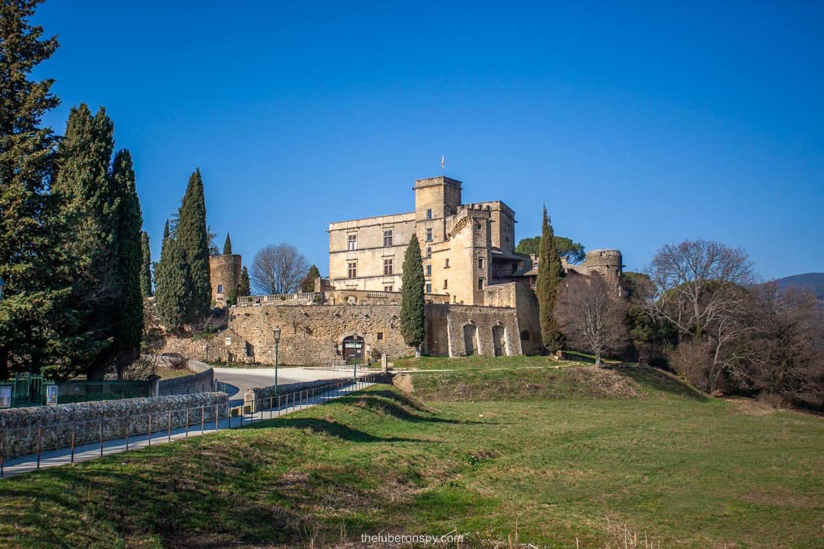 The Château de Lourmarin, a historic Renaissance castle in Provence, France, stands surrounded by cypress trees and rolling greenery under a clear blue sky. Its fortified stone walls and medieval towers contrast beautifully with the tranquil landscape.