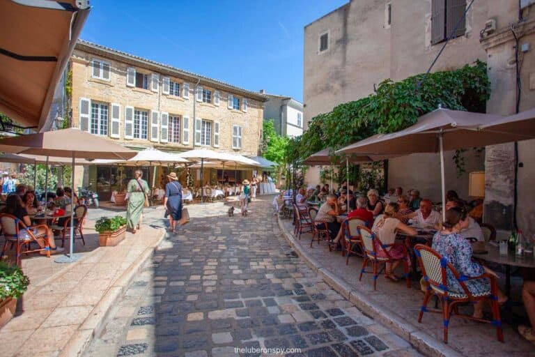 A picturesque cobblestone street in Lourmarin, lined with lively outdoor cafés shaded by large umbrellas. People enjoy meals and conversations at classic French bistro tables, while others stroll past charming stone buildings with shuttered windows.