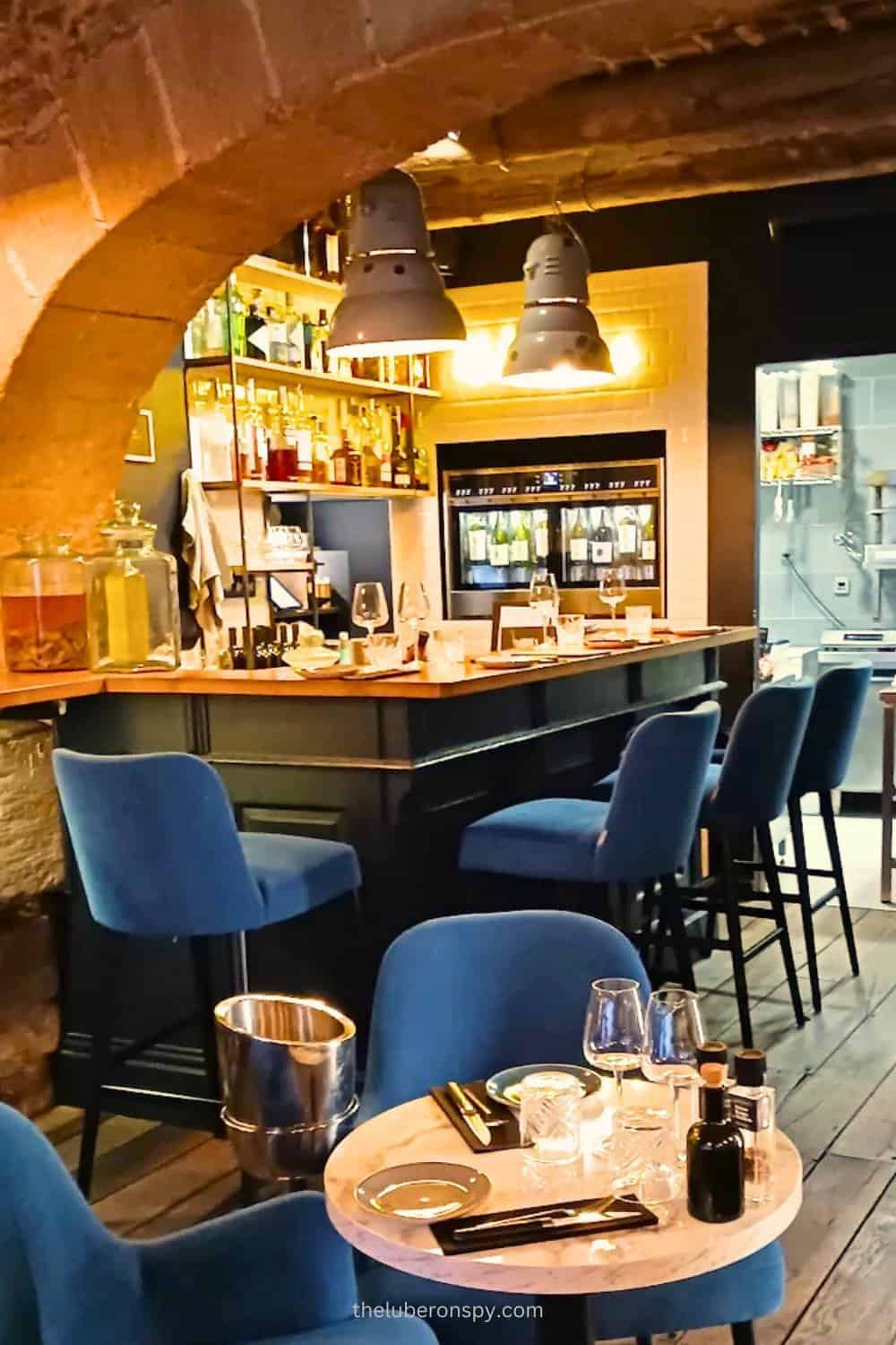 Cozy interior of a modern bar and dining area at Bouchon in Lourmarin, featuring a marble-topped table, blue velvet chairs, and a well-stocked wine and spirits bar under warm lighting.