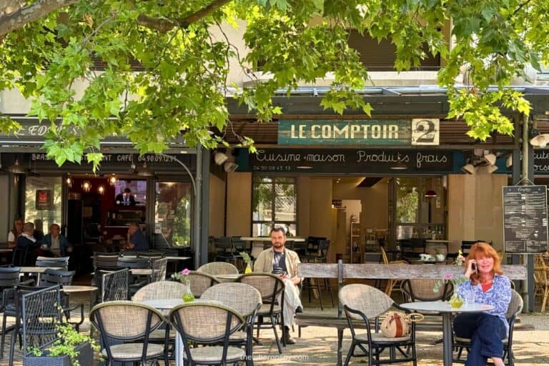 Outdoor seating in front of Le Comptoir café in Lourmarin, with a few people relaxing under leafy trees and a rustic sign reading “Cuisine maison, Produits frais” above the entrance.