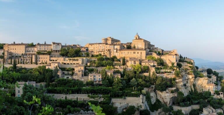 Panoramic view of Gordes, a hilltop village in Provence, with layers of historic stone buildings, lush gardens, and dramatic cliffs under a soft evening sky.