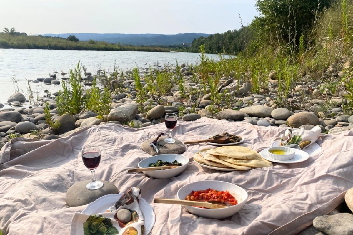Picnic setup on a rocky riverbank with dishes of flatbread, sautéed greens, tomato stew, olive oil, and glasses of red wine laid out on a beige blanket, with a scenic view of the river and distant hills.