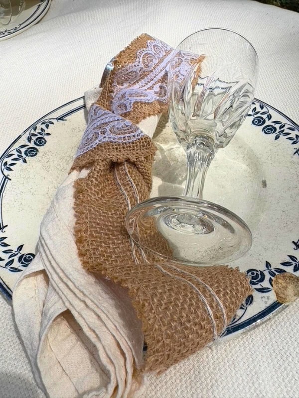 Close-up of a rustic table setting with a clear wine glass resting on a vintage-style plate, paired with a cloth napkin wrapped in burlap and lace.