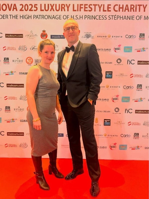 The Luberon chauffeur is in a black tuxedo and bow tie stands on a red carpet alongside a woman in a fitted sleeveless dress, posing in front of a white backdrop with logos and the header “Luxury Lifestyle Charity” under the high patronage of a Monaco royal.