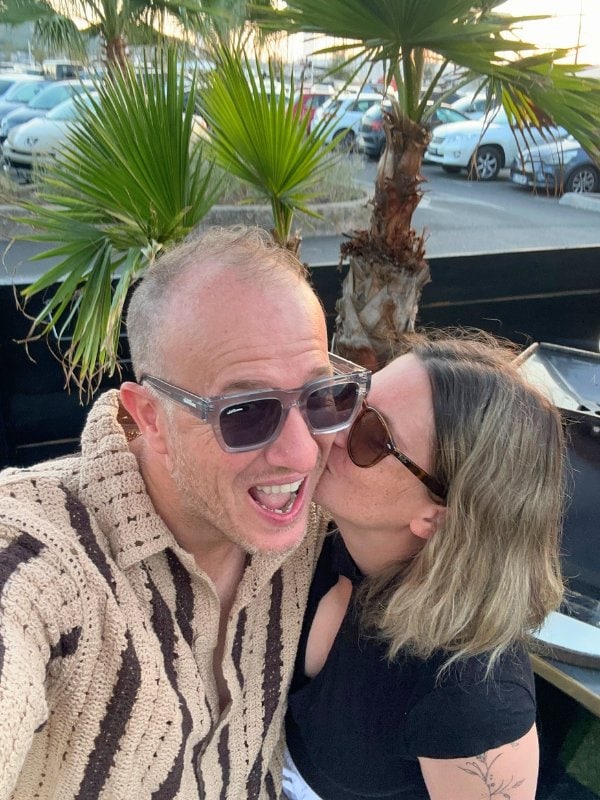 A man in sunglasses and a crochet-style striped shirt smiles widely while a woman kisses his cheek.