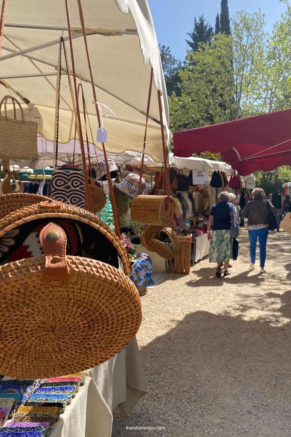 Woven straw bags and colorful beaded accessories hang at a busy Lourmarin market beneath white and red market canopies