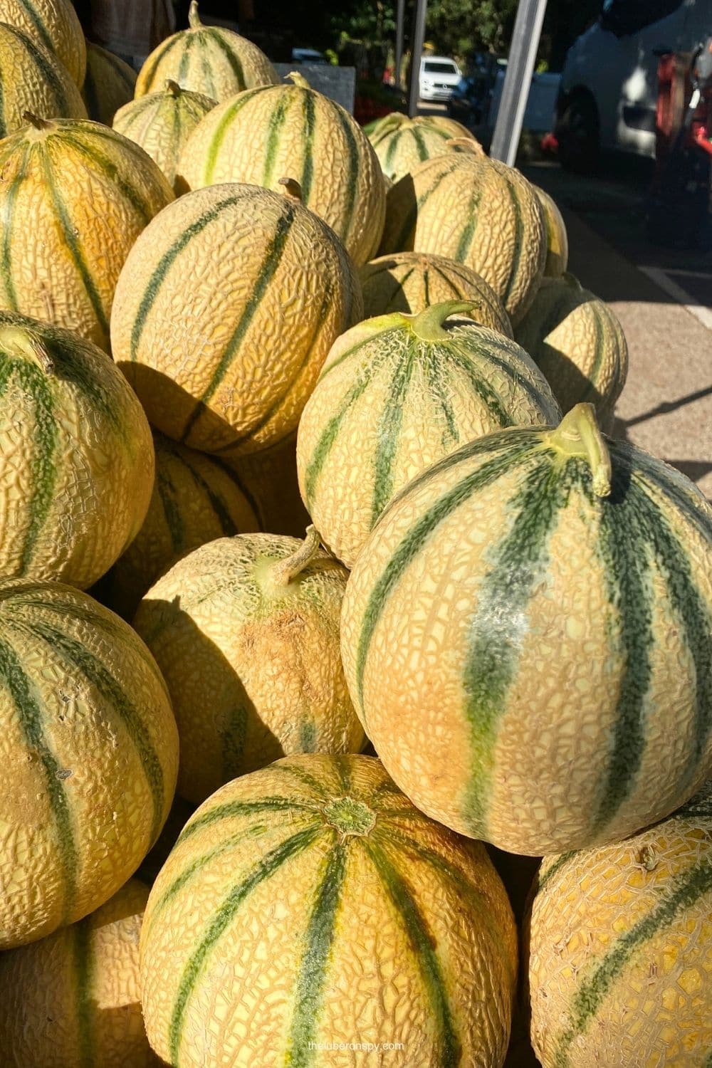 A large pile of ripe cantaloupes with pale orange skin and green stripes fills a produce stall at the Lourmarin market.