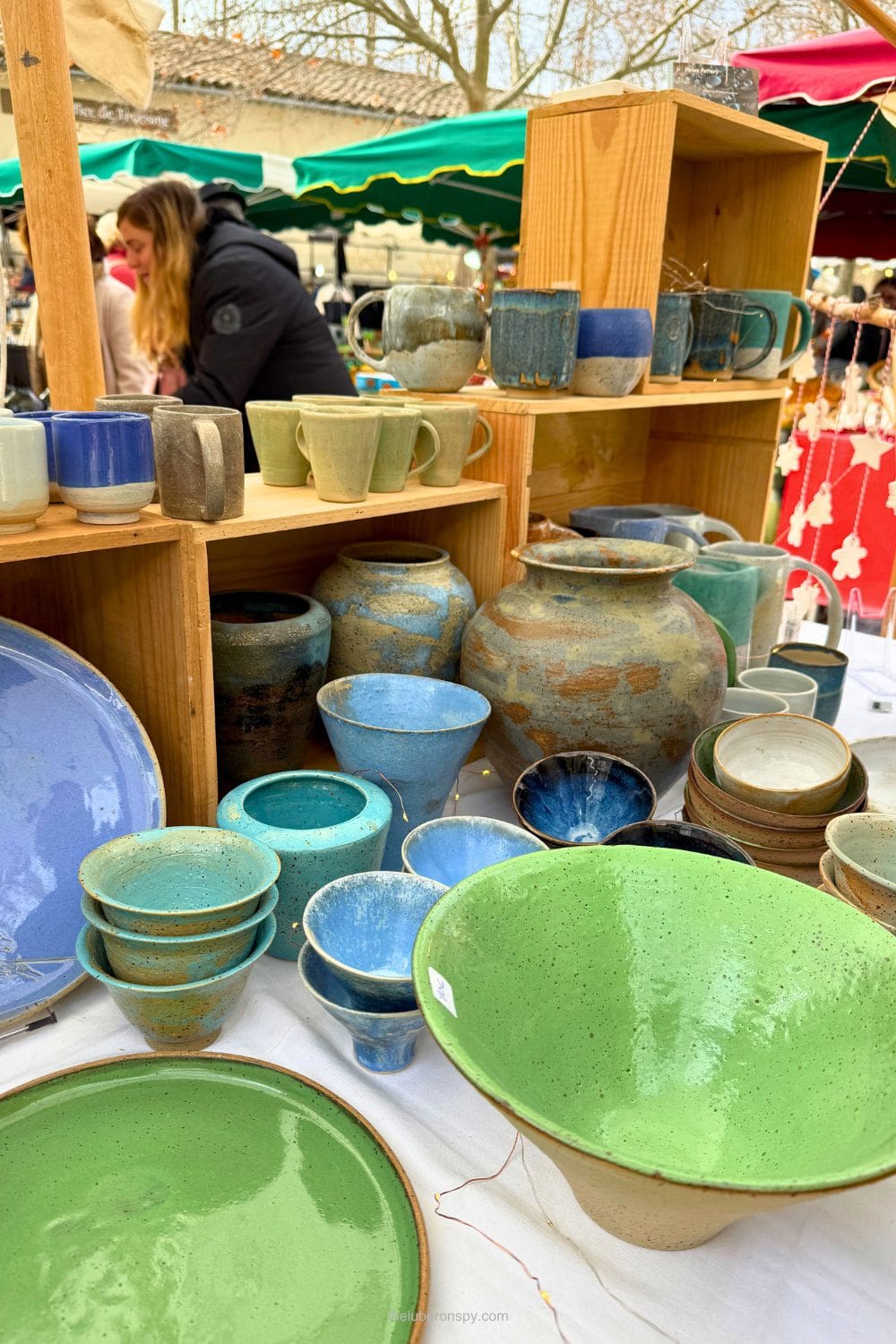 Handcrafted ceramic cups, bowls, vases, and plates in blue, green, and earthy tones displayed at a lively Provence market.