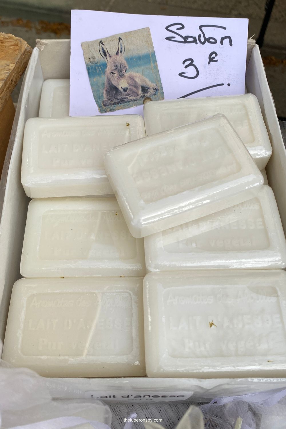 White donkey milk soap bars stacked in a market display with a handwritten Savon 3โฌ sign and a small donkey illustration.