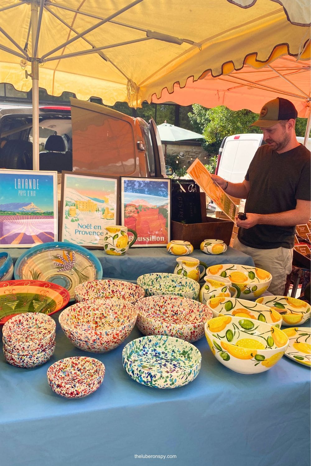 A Lourmarin market stall displays colorful hand-painted ceramic bowls and lemon-patterned dishes beneath Luberon posters.