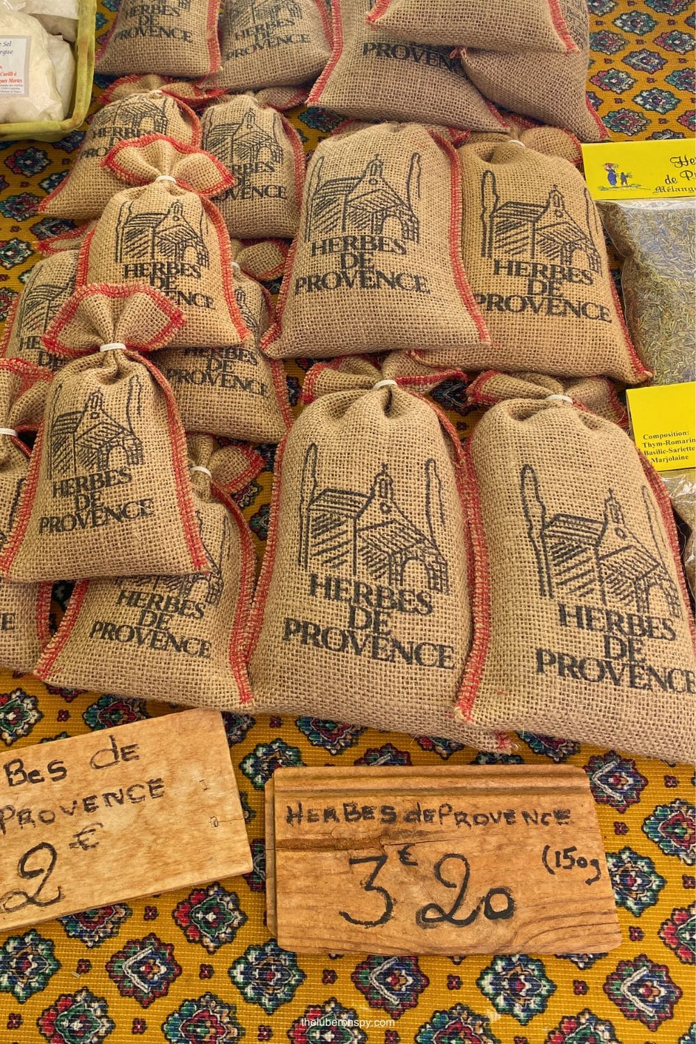 Burlap bags of Provenรงal herbs are displayed at the Lourmarin market with labels reading HERBES DE PROVENCE and signs showing 2โฌ and โฌ3.20.