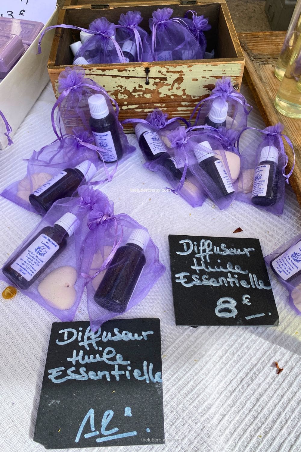 Small bottles of essential oils are arranged in purple organza bags around a rustic wooden box beneath signs reading "Diffuseur Huile Essentielle."