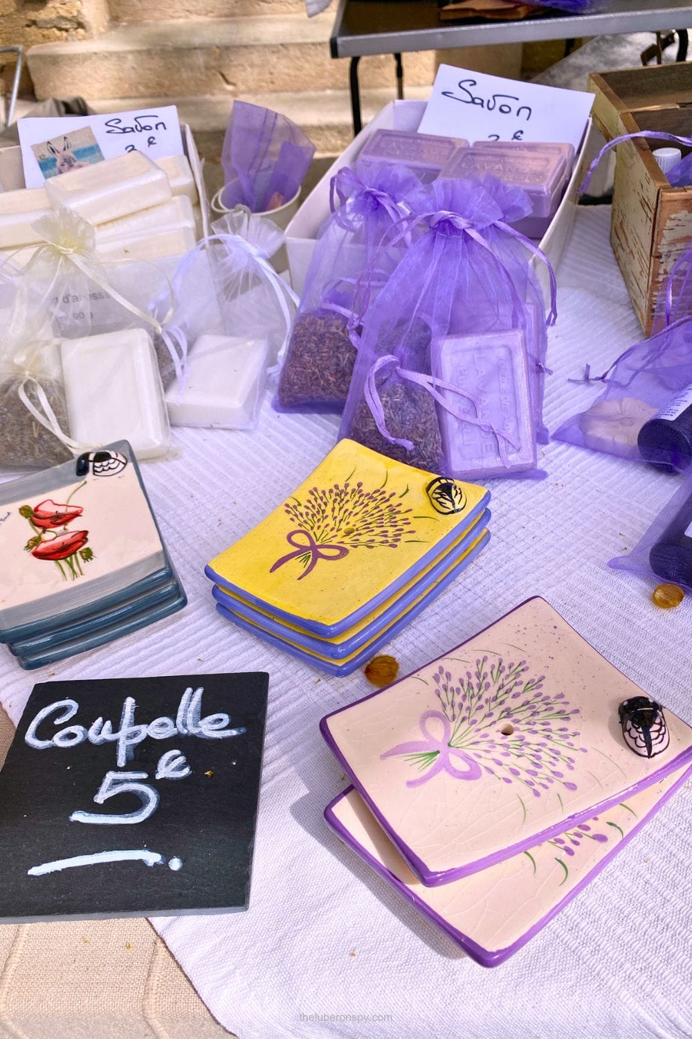A market table displays handmade white and purple soaps, lavender sachets, and ceramic dishes beneath signs reading Savon and Coupelle 5โฌ.