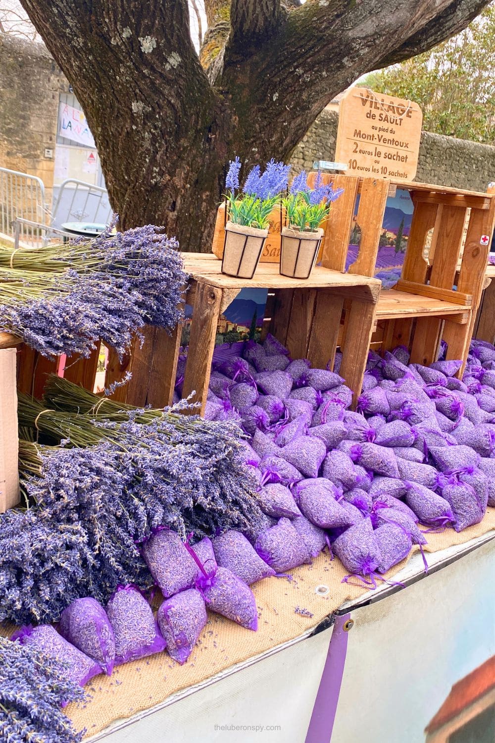 Bundles of dried lavender and purple lavender sachets fill a market stall beneath a sign reading "VILLAGE de SAULT au pied du Mont Ventoux" and "2 euros le sachet ou 10 euros les 10 sachets."