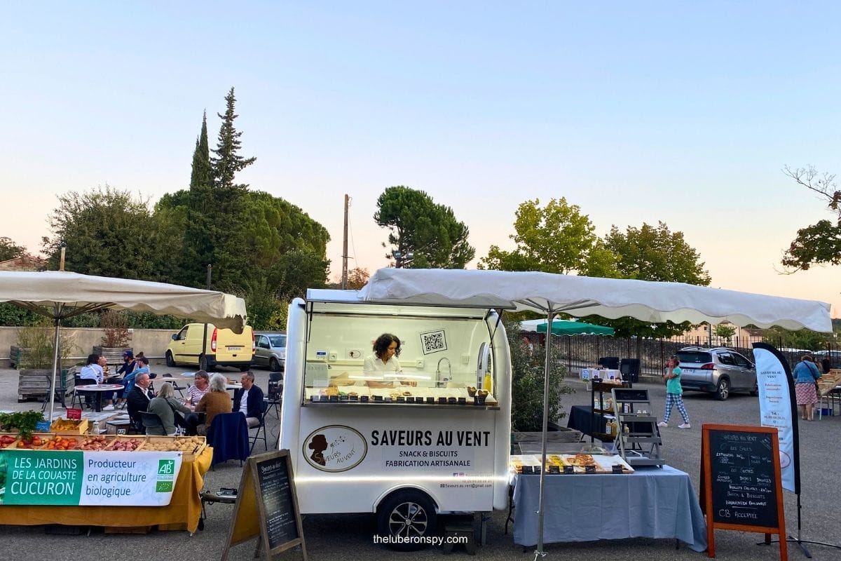 A Luberon Provence market with a food truck selling handmade snacks and biscuits alongside local produce and artisan stalls.