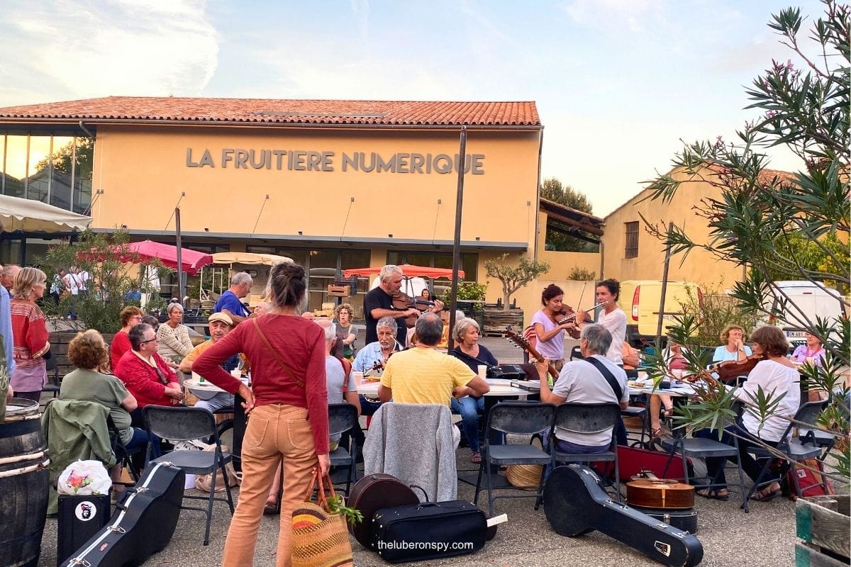 Musicians playing guitars and other instruments as people sit together outdoors near La Fruitiรจre Numรฉrique in Lourmarin.