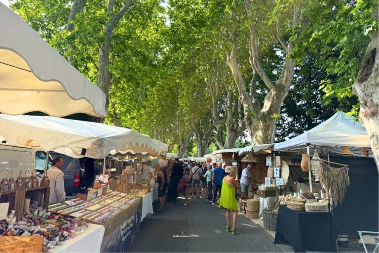 A busy Lourmarin market in Provence lined with white-canopy stalls selling handmade crafts, woven baskets, textiles, and local goods beneath a canopy of leafy plane trees.