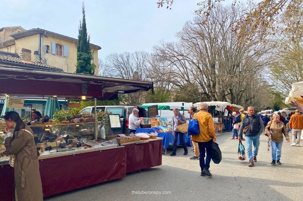 A bustling Lourmarin market with shoppers browsing food and clothing stalls along a village street lined with leafless plane trees.