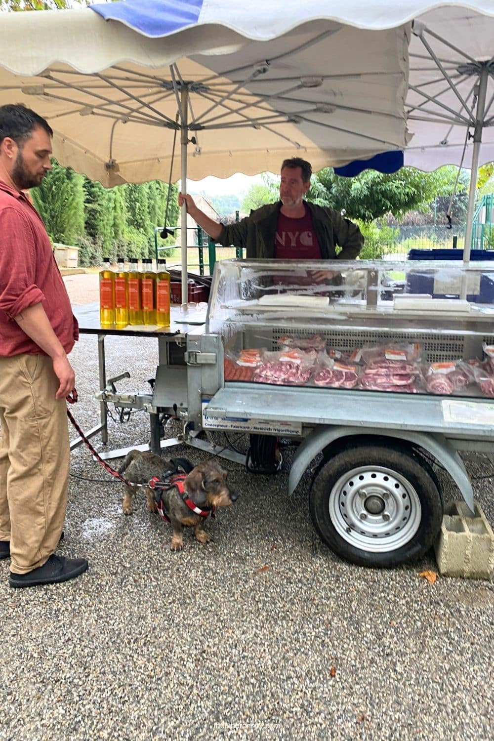 A vendor sells fresh lamb meat and bottles of olive oil, with a shopper and small dog nearby.