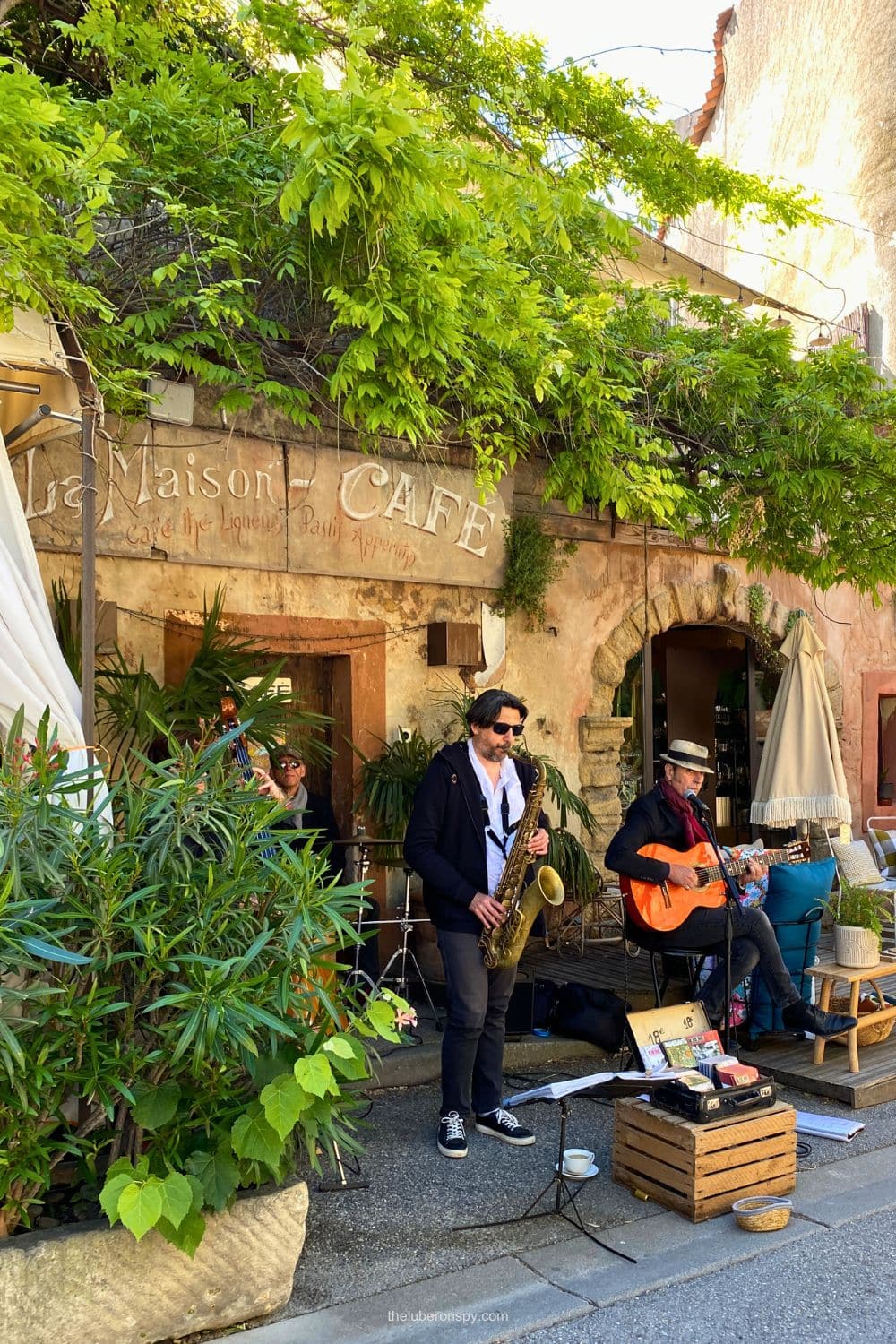A saxophonist and guitarist perform outside La Maison Cafรฉ during a lively market day in Lourmarin.