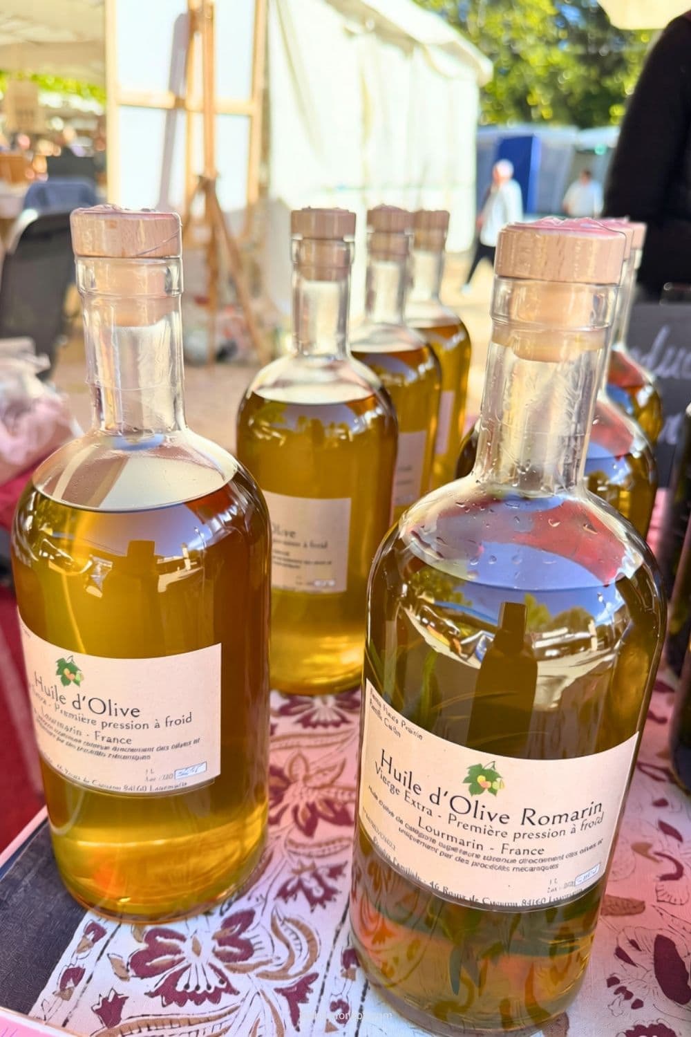 Bottles of olive oil labeled "Huile d'Olive Romarin", "Vierge Extra", "Premiรจre pression ร froid", and "Lourmarin - France" are displayed at the market.