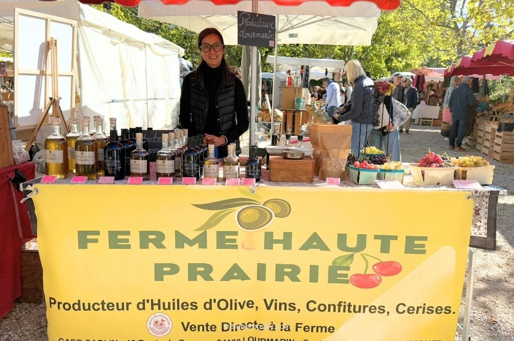 A producer sells olive oil, wine, jams, and cherries at the Lourmarin market beneath a sign reading "FERME HAUTE PRAIRIE", "Producteur d'Huiles d'Olive, Vins, Confitures, Cerises", and "Vente Directe ร la Ferme".