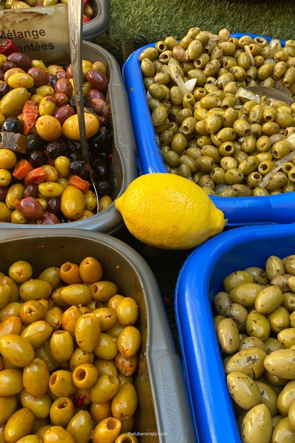 Trays of green and mixed olives surround a bright yellow lemon at a Lourmarin market stall.