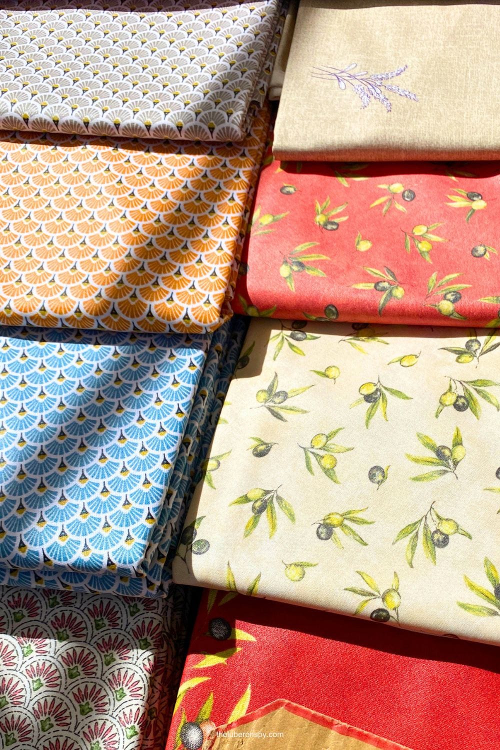 Stacks of colorful Provenรงal tablecloths featuring olive branches, lemons, lavender sprigs, and geometric patterns at a lavender Provence market.