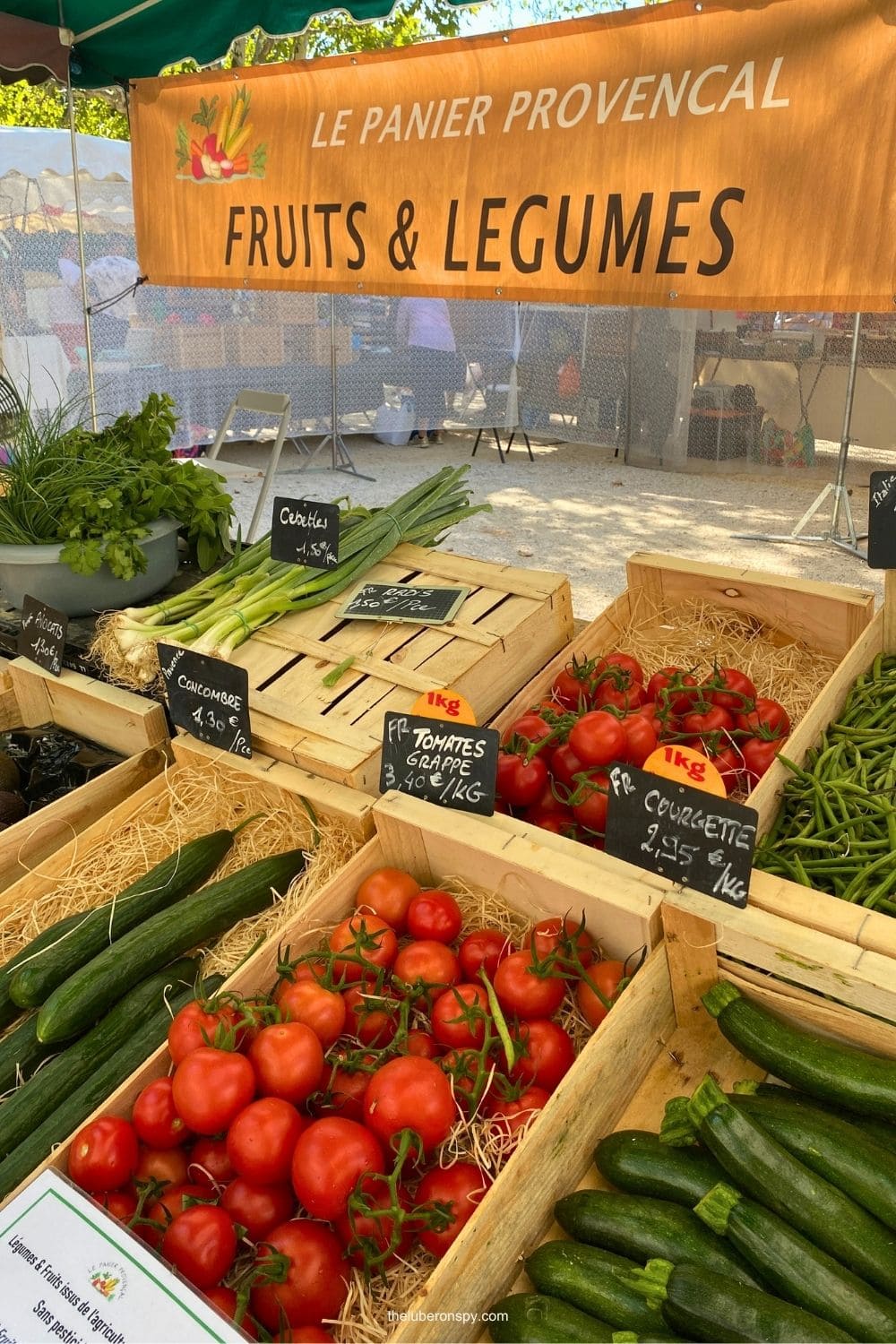 A colorful Lourmarin market stall displays fresh Provenรงal fruits and vegetables including tomatoes, cucumbers, spring onions, and herbs beneath a sign reading "LE PANIER PROVENCAL" and "FRUITS & LEGUMES".